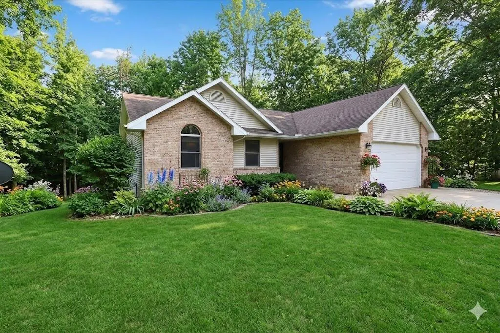 A tan brick house with a brown roof, white garage door, and green lawn is surrounded by trees and flowers.
