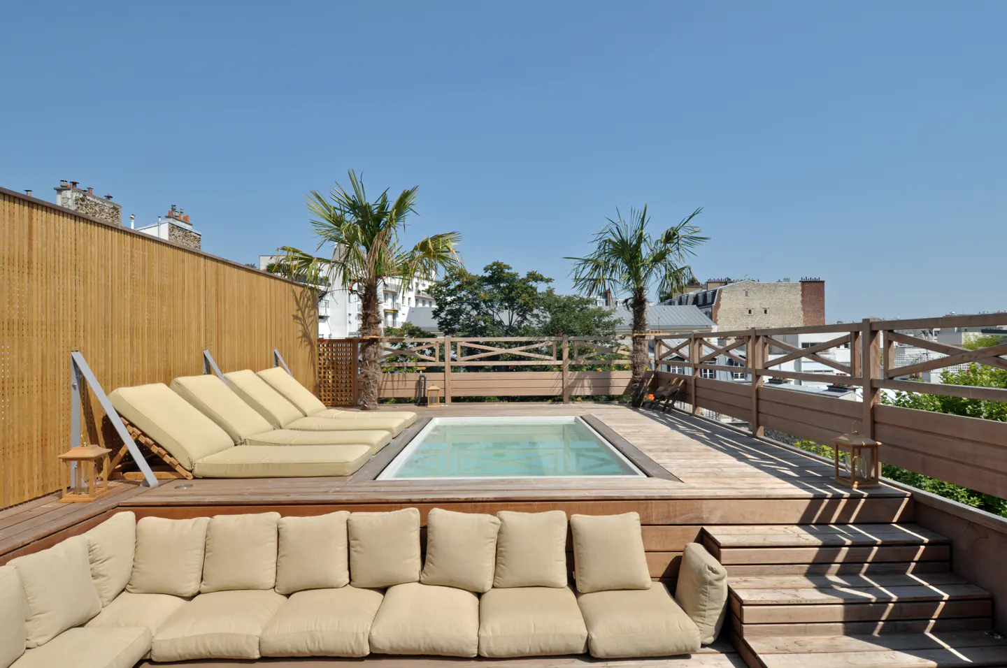 Rooftop deck with a small pool, lounge chairs, and a beige sectional sofa. Palm trees and a wooden fence surround the deck.