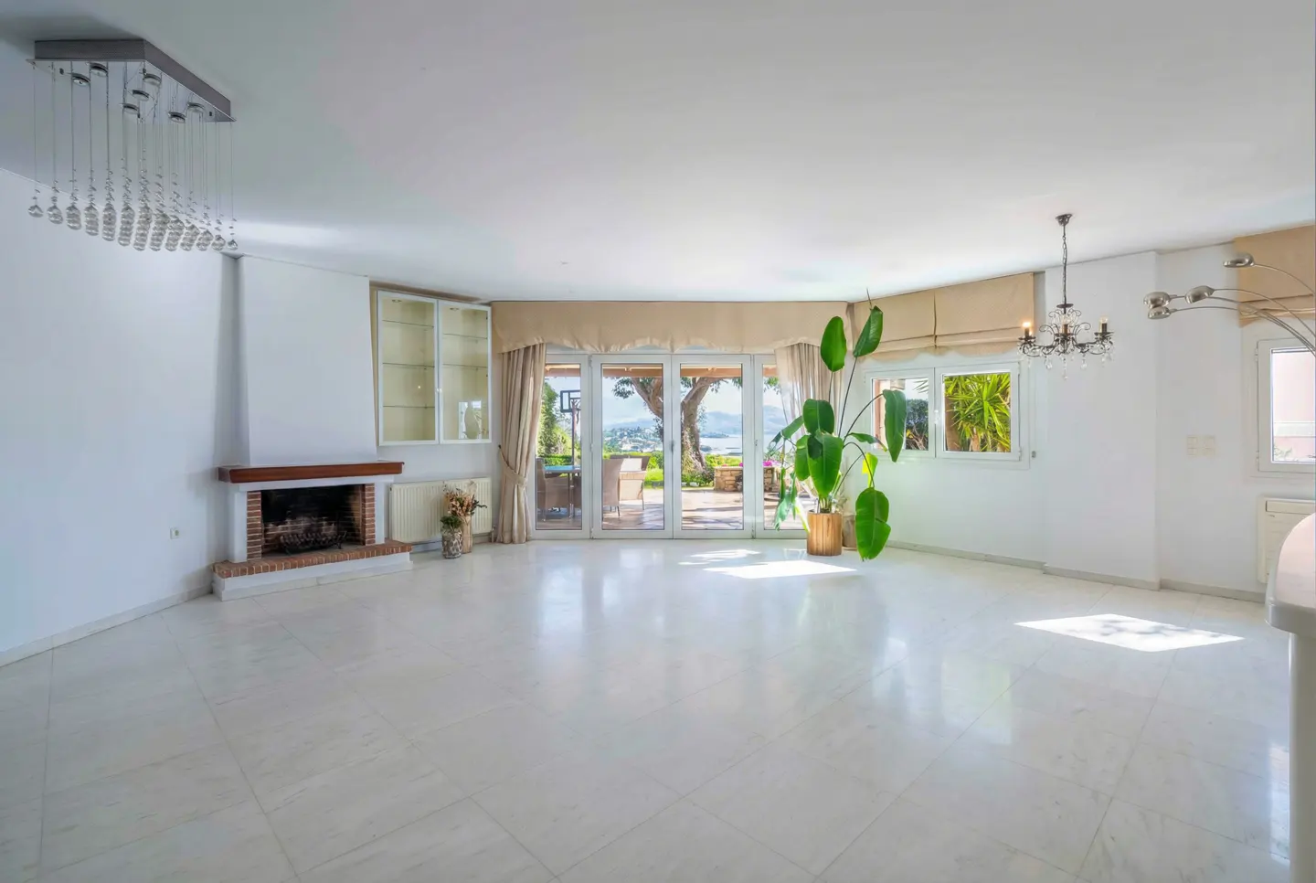 Bright, empty living room with white marble floors, fireplace, and glass doors to a patio with an ocean view. Chandeliers hang from the ceiling.