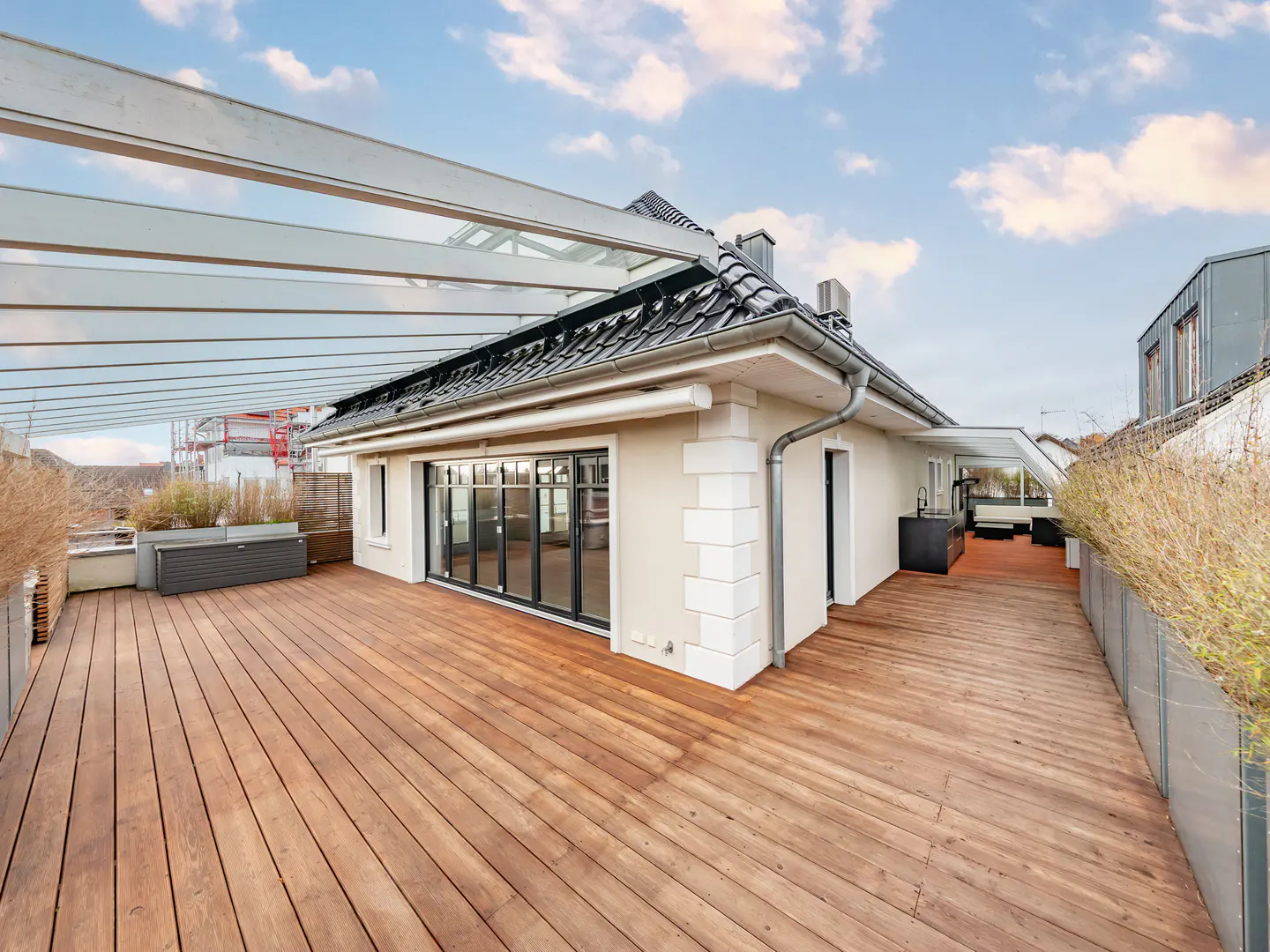 Wide shot of a rooftop deck with wood flooring, a white house with black trim, and a metal pergola against a blue sky.