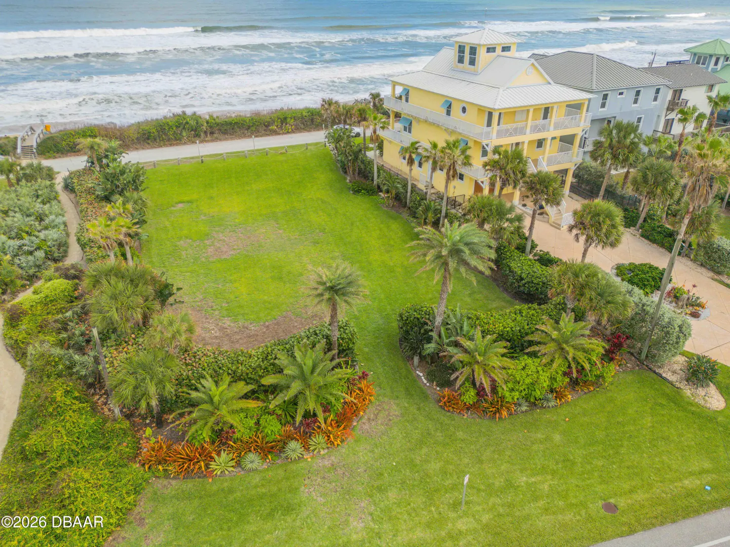 Aerial view of a yellow, multi-story beach house with a large lawn and palm trees, near the ocean.