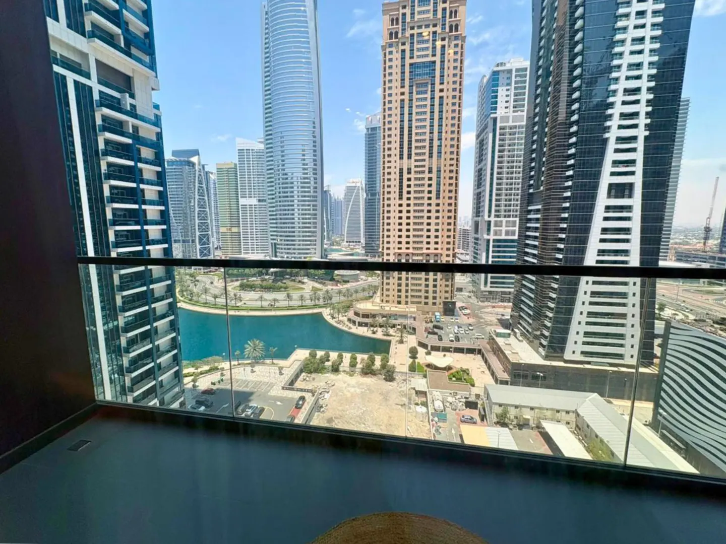 View from a high-rise balcony with glass railings overlooking a blue canal and city skyscrapers on a sunny day.