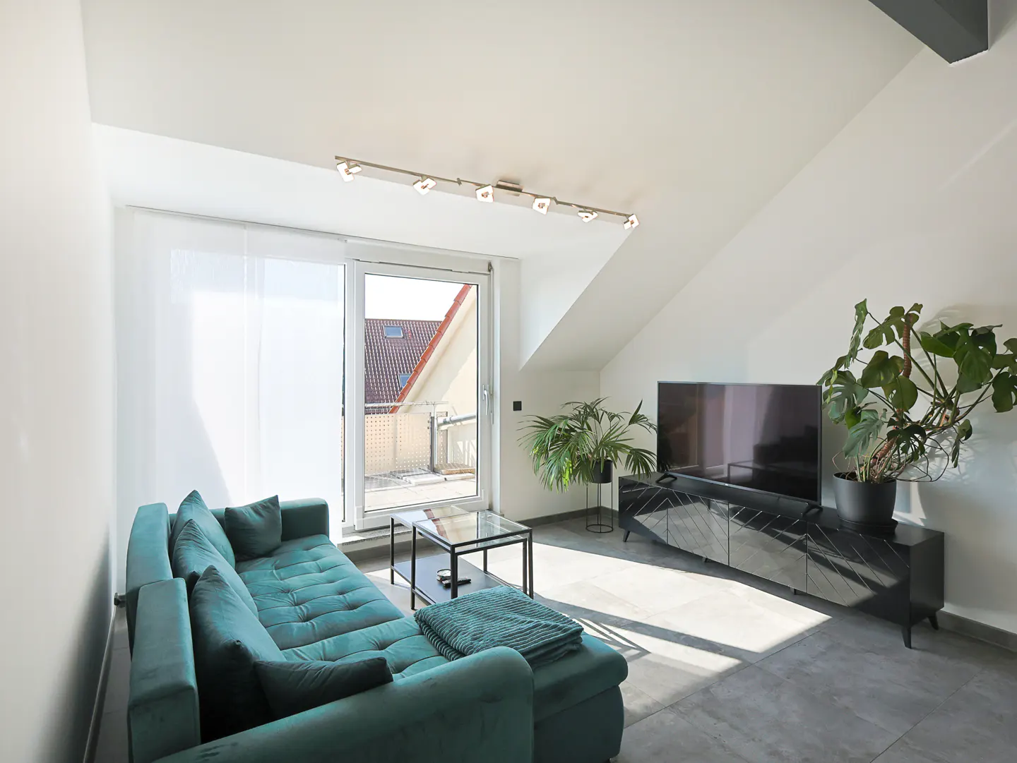 Bright living room with a teal sofa, glass table, TV, and plants. Sunlight streams in from a sliding glass door.
