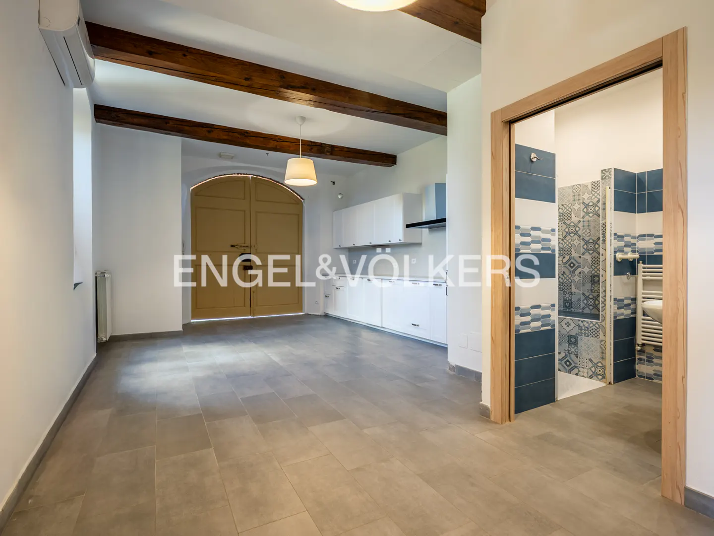 An interior shot of a modern kitchen with white cabinets, a large wooden door, and a bathroom with blue and white tiles.