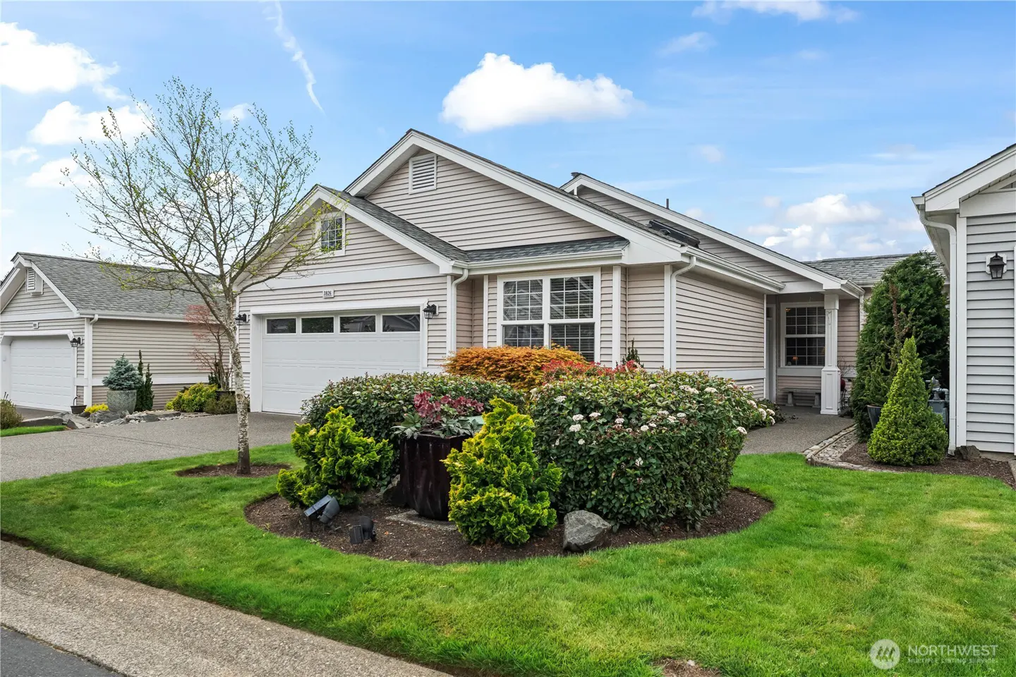 Beige siding house with white garage door, green lawn, and flower bed. Blue sky with clouds.