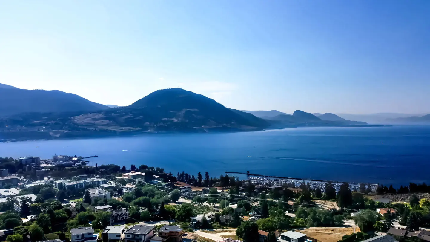 Aerial view of a blue lake surrounded by lush green hills and a coastal town with a marina under a clear blue sky.