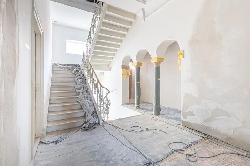 A bright, white hallway under renovation with stairs, columns, and exposed wires on the floor.