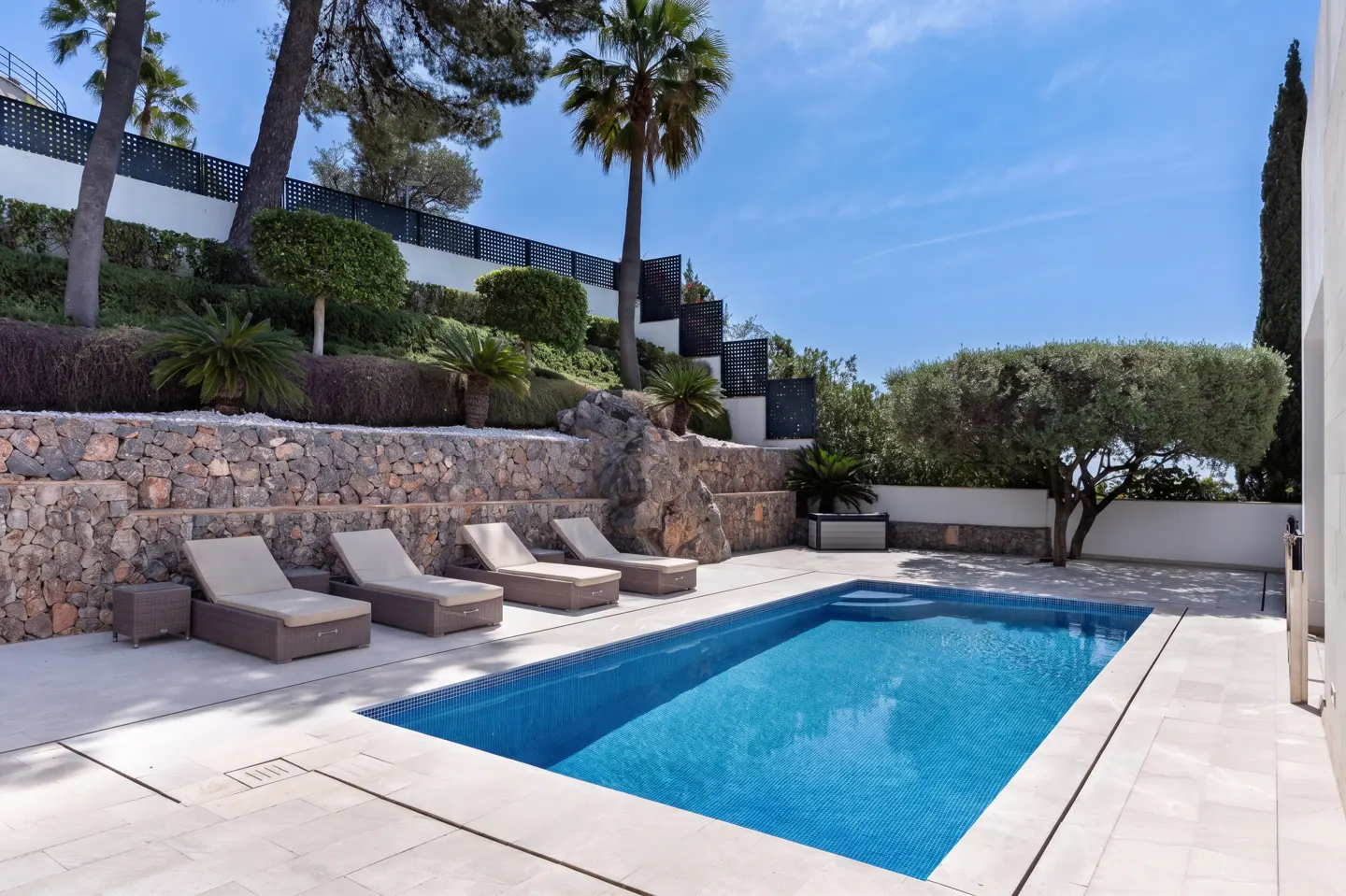 A backyard pool with blue tiles is surrounded by lounge chairs and a stone wall with greenery.
