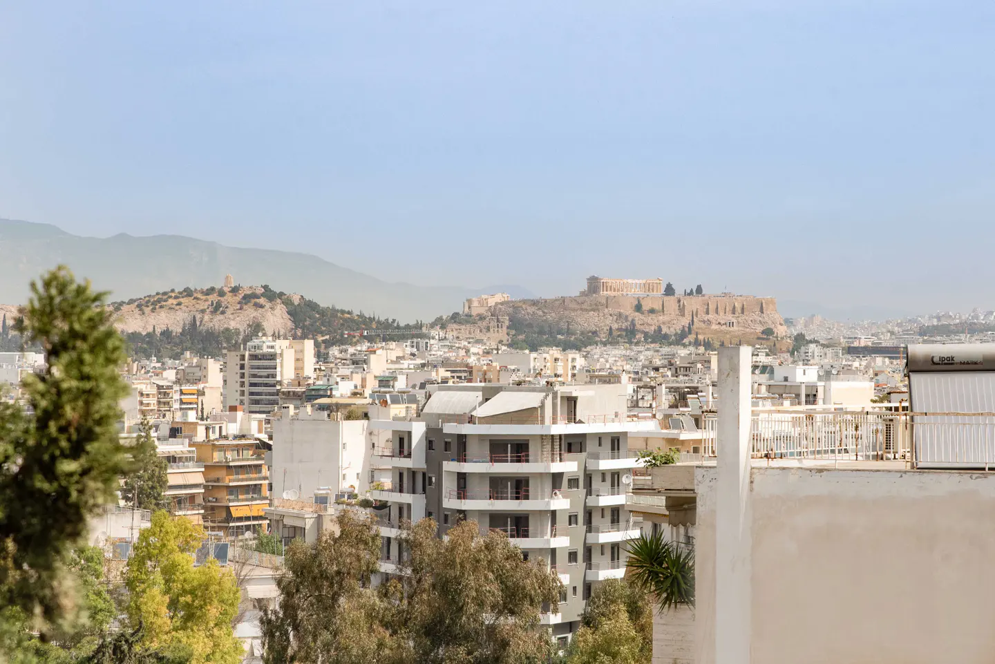 Athens cityscape view with the Acropolis in the background on a hazy day. Buildings and trees in the foreground.
