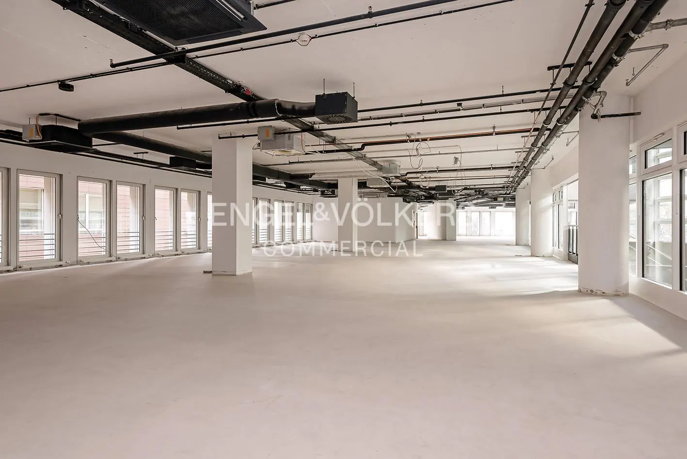 Bright, empty commercial space with white walls, pillars, and floor. Black pipes and vents run across the ceiling. Large windows line the walls.