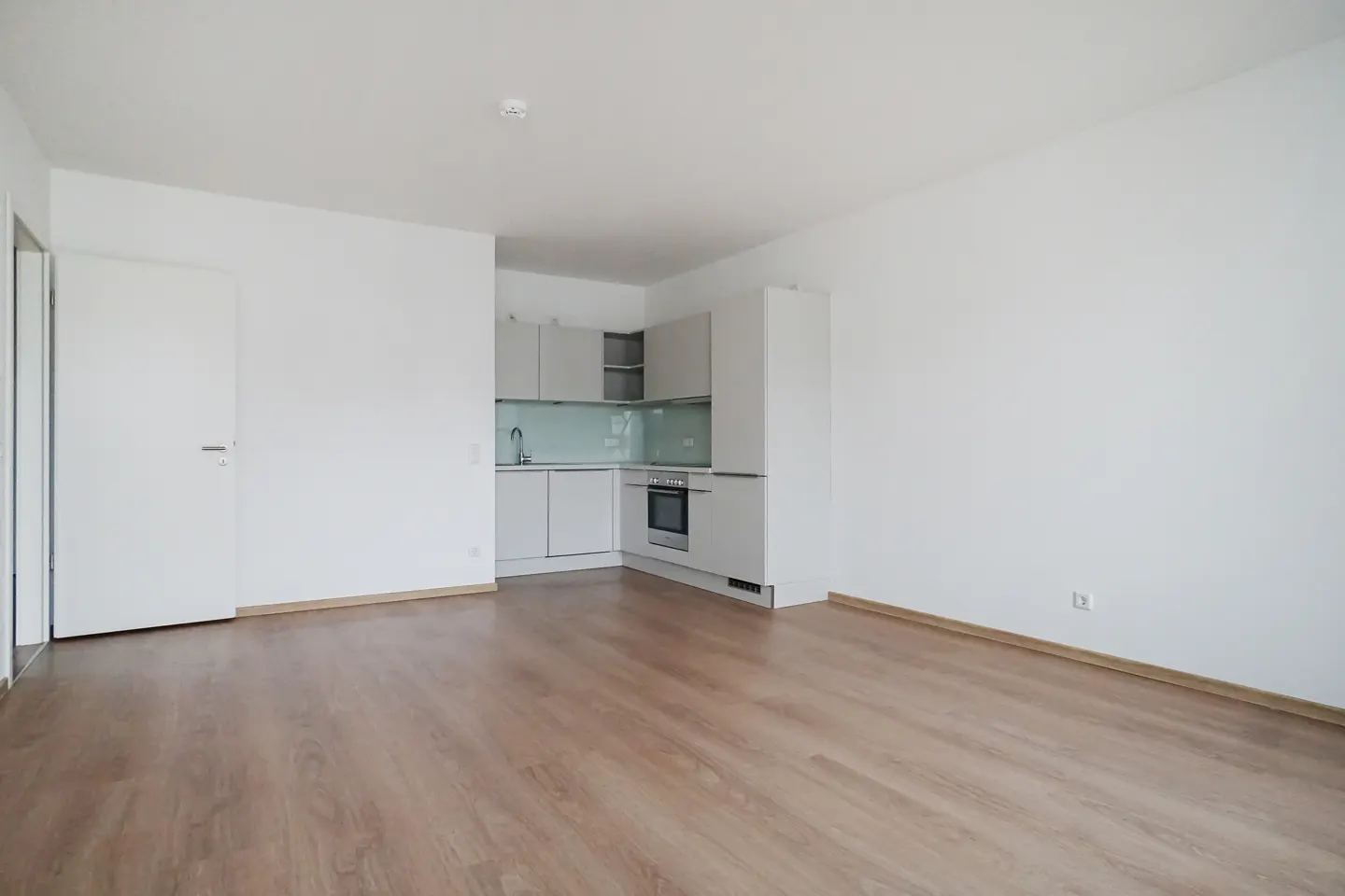 Bright, empty apartment with wood floors, white walls, and a modern kitchen with light gray cabinets. A white door is on the left.