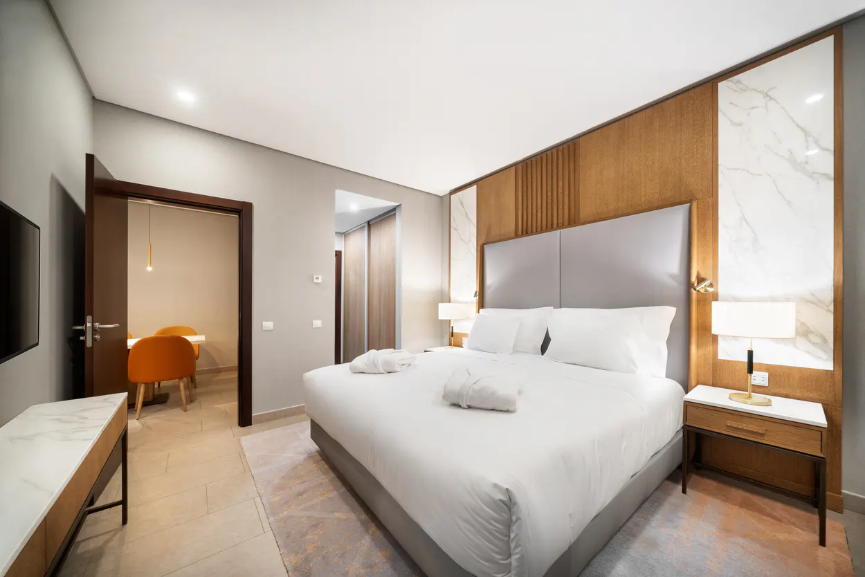 A modern hotel room features a king bed with white linens and robes, a wood-paneled accent wall, and a glimpse into a dining area.