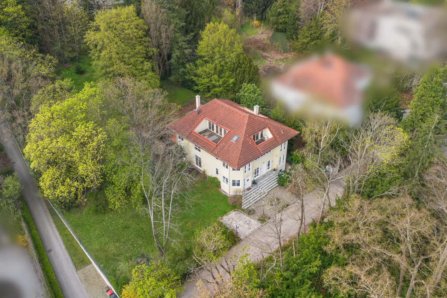 Aerial view of a cream-colored house with a red tile roof, surrounded by lush green trees and a paved driveway.