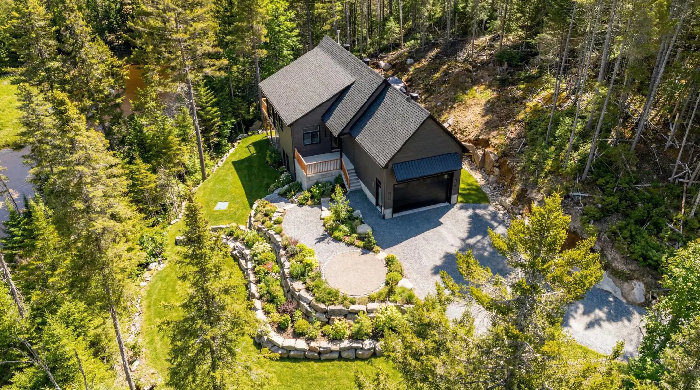 Aerial view of a modern, dark gray house with a black roof, surrounded by lush green trees and landscaping.