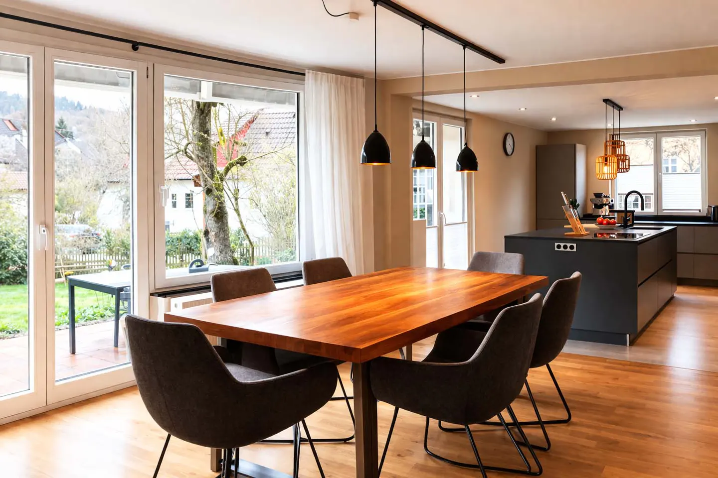 Bright dining area with a wooden table, six gray chairs, and black pendant lights. Large windows overlook a green yard and a modern kitchen is visible in the background.