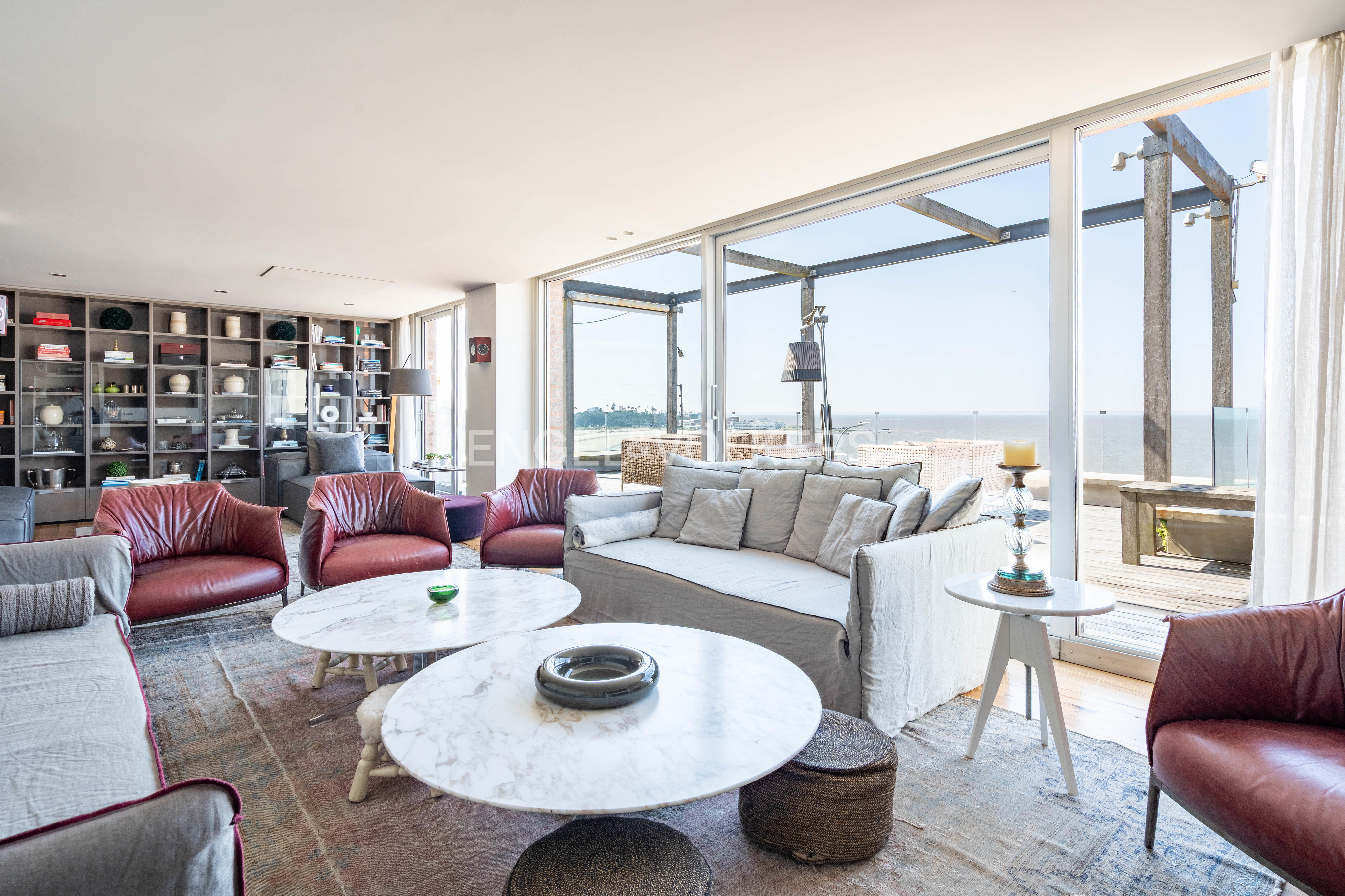 Modern living room with red leather armchairs, a grey sofa, two marble coffee tables, and floor-to-ceiling ocean view windows. [AI-generated text]