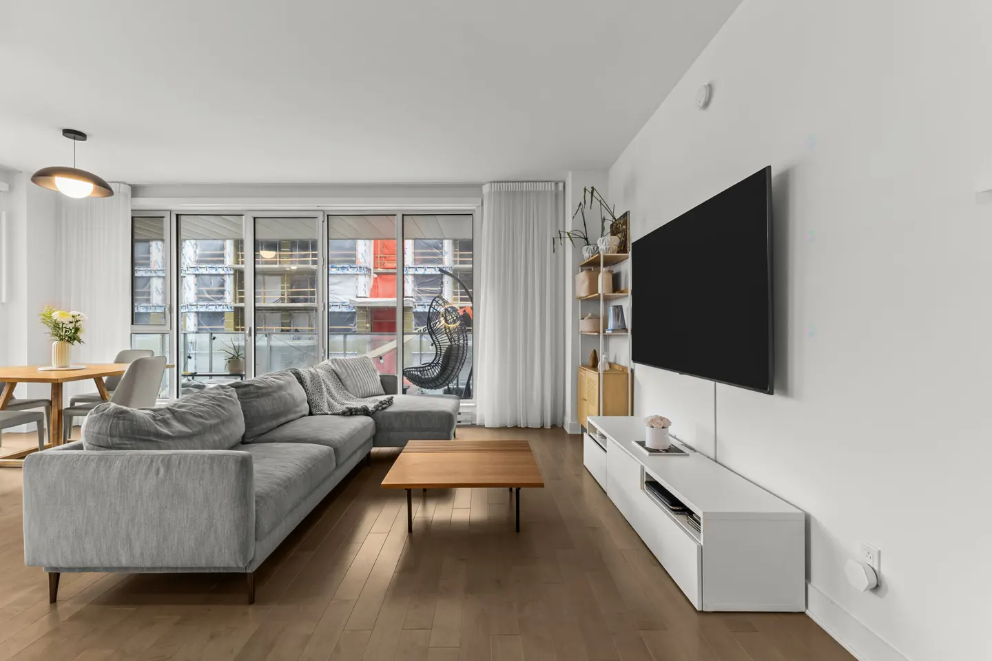 Bright living room with a gray sectional sofa, wood floors, a TV, and a dining table near a window.
