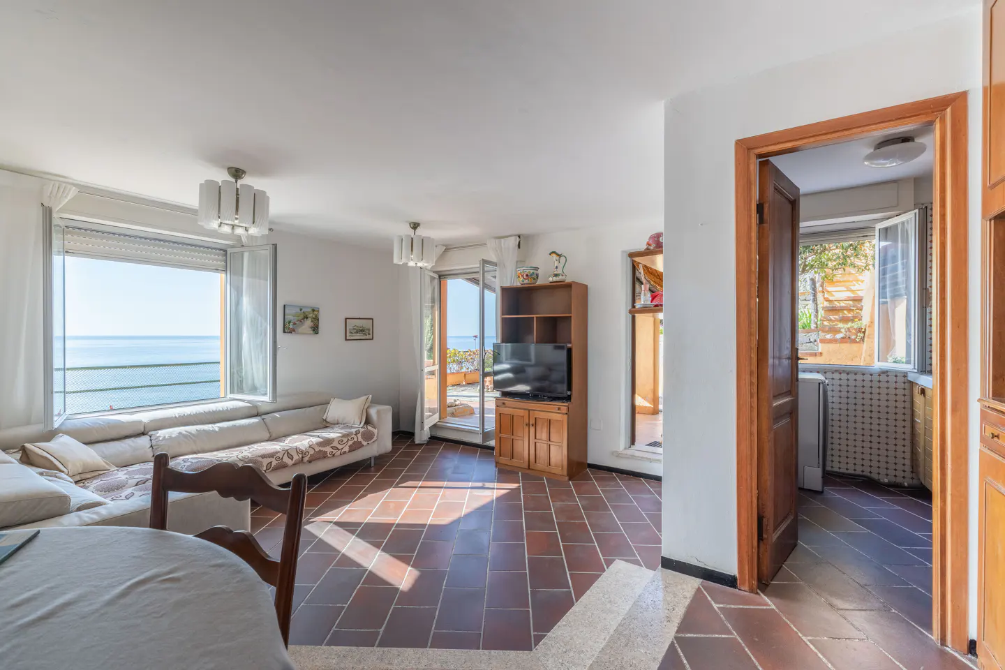 Bright living room with a white sofa, brown tile floor, and a large window overlooking the ocean. A wooden cabinet and doorway are visible.