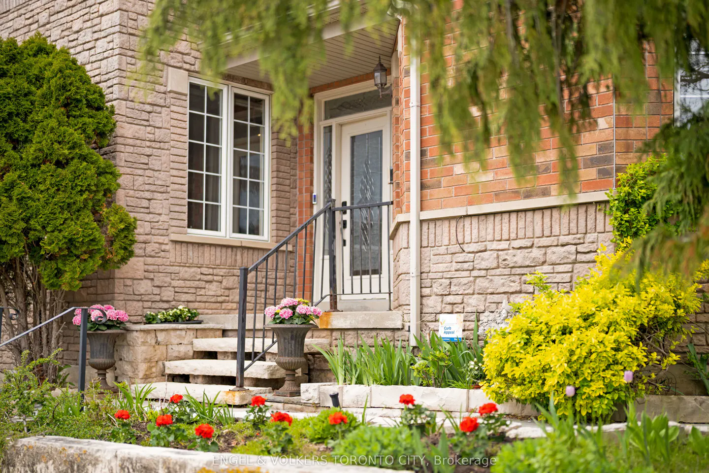 Exterior of a two-story brick house with stone accents, a white front door, and a black railing leading to the entrance. Red and yellow flowers in the garden.