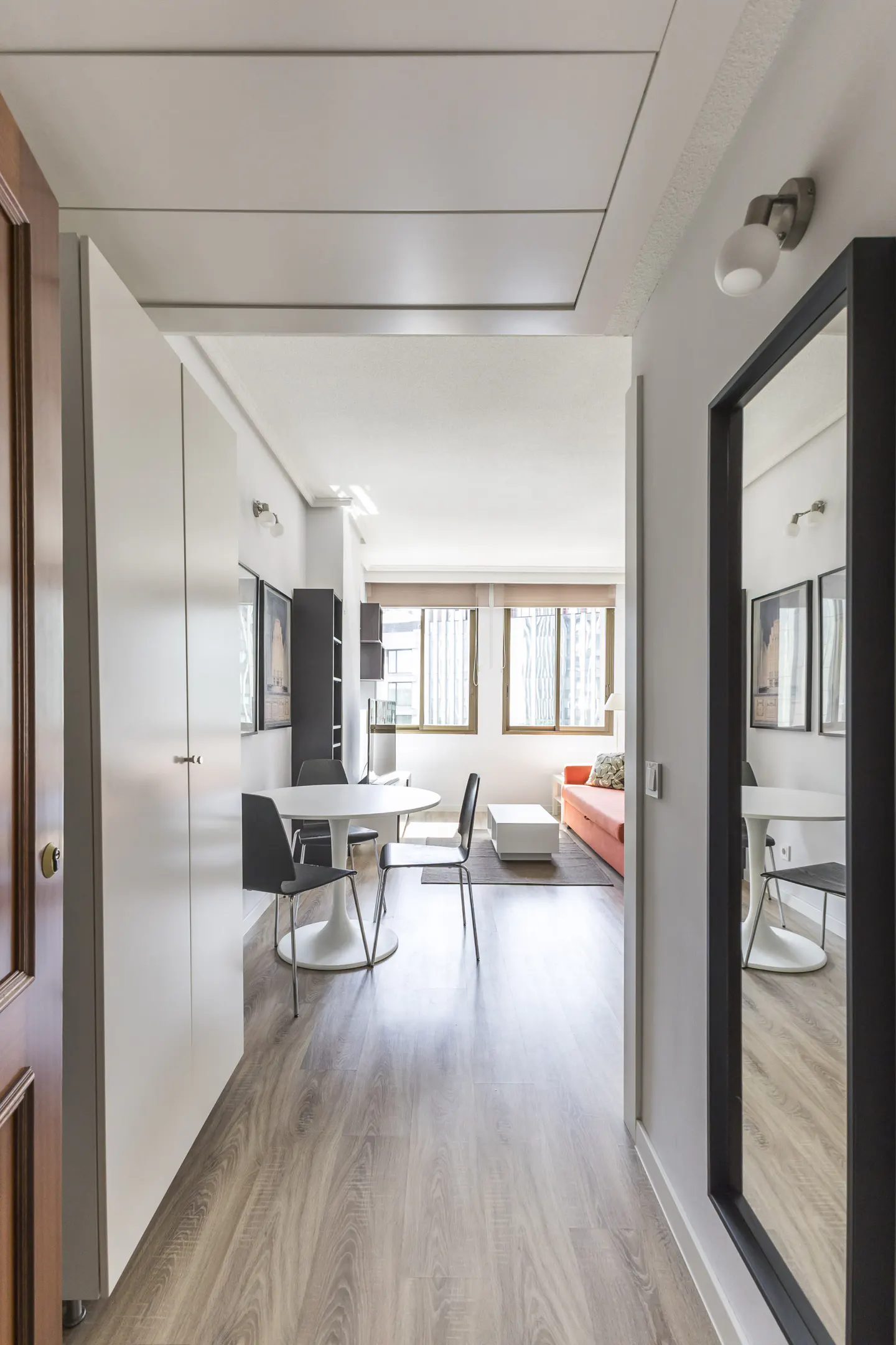 Hallway view into a modern apartment with light wood floors, white walls, and a round white table with black chairs. A mirror reflects the scene.