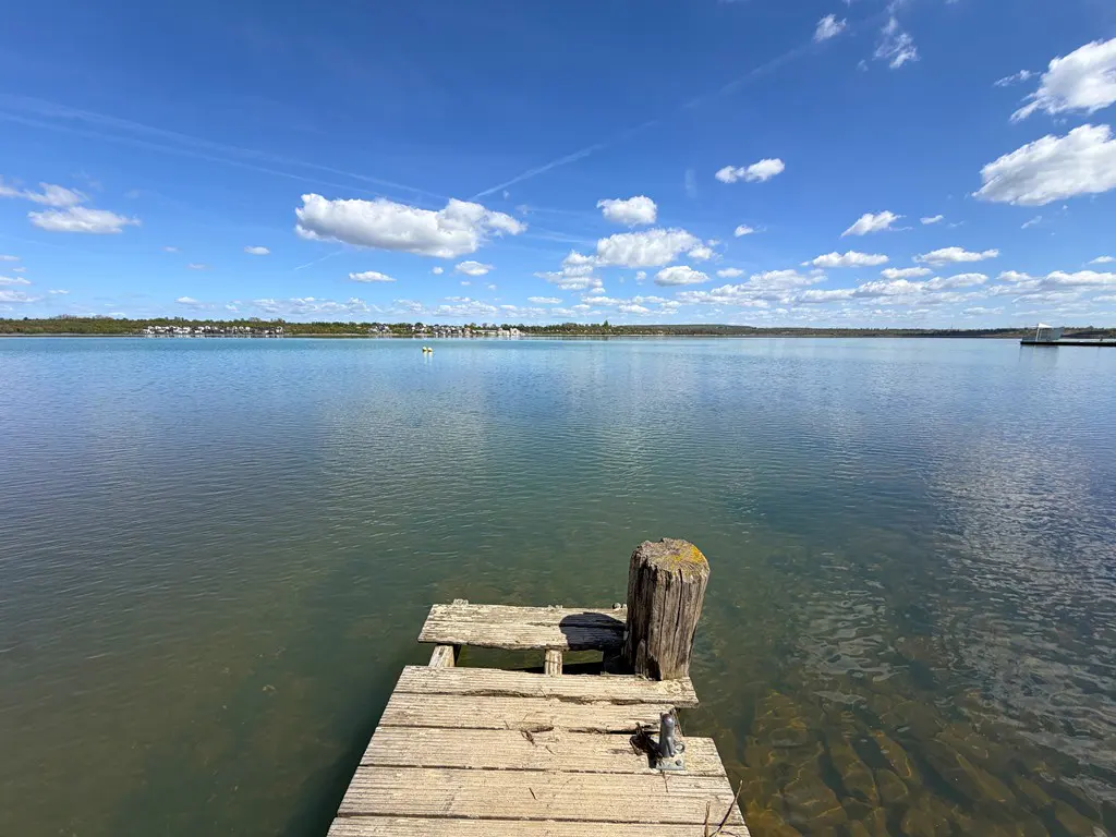 A weathered wooden dock extends into a calm lake under a blue sky with scattered clouds. Distant shoreline with houses visible.