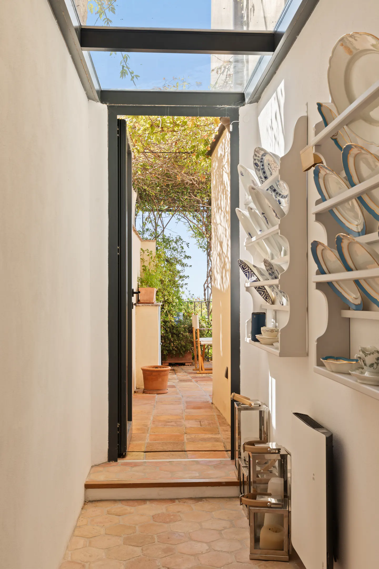 Indoor hallway with terracotta tile floor, white walls, and a glass ceiling. An open door leads to a patio with greenery and a table. Decorative plates are displayed on shelves.