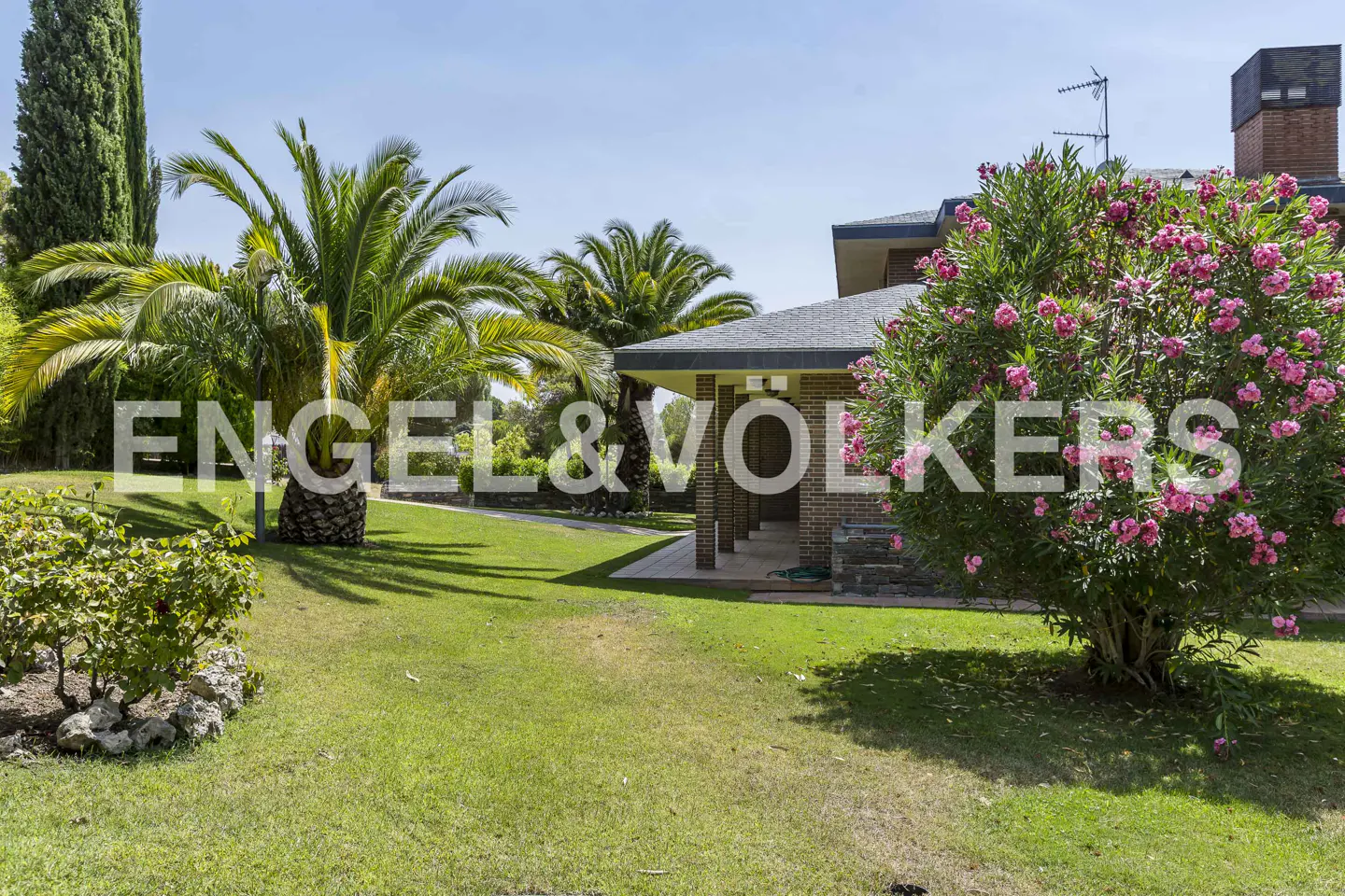 Exterior view of a brick house with a gray roof, surrounded by palm trees, green grass, and pink flowers. The Engel & Volkers logo is superimposed.