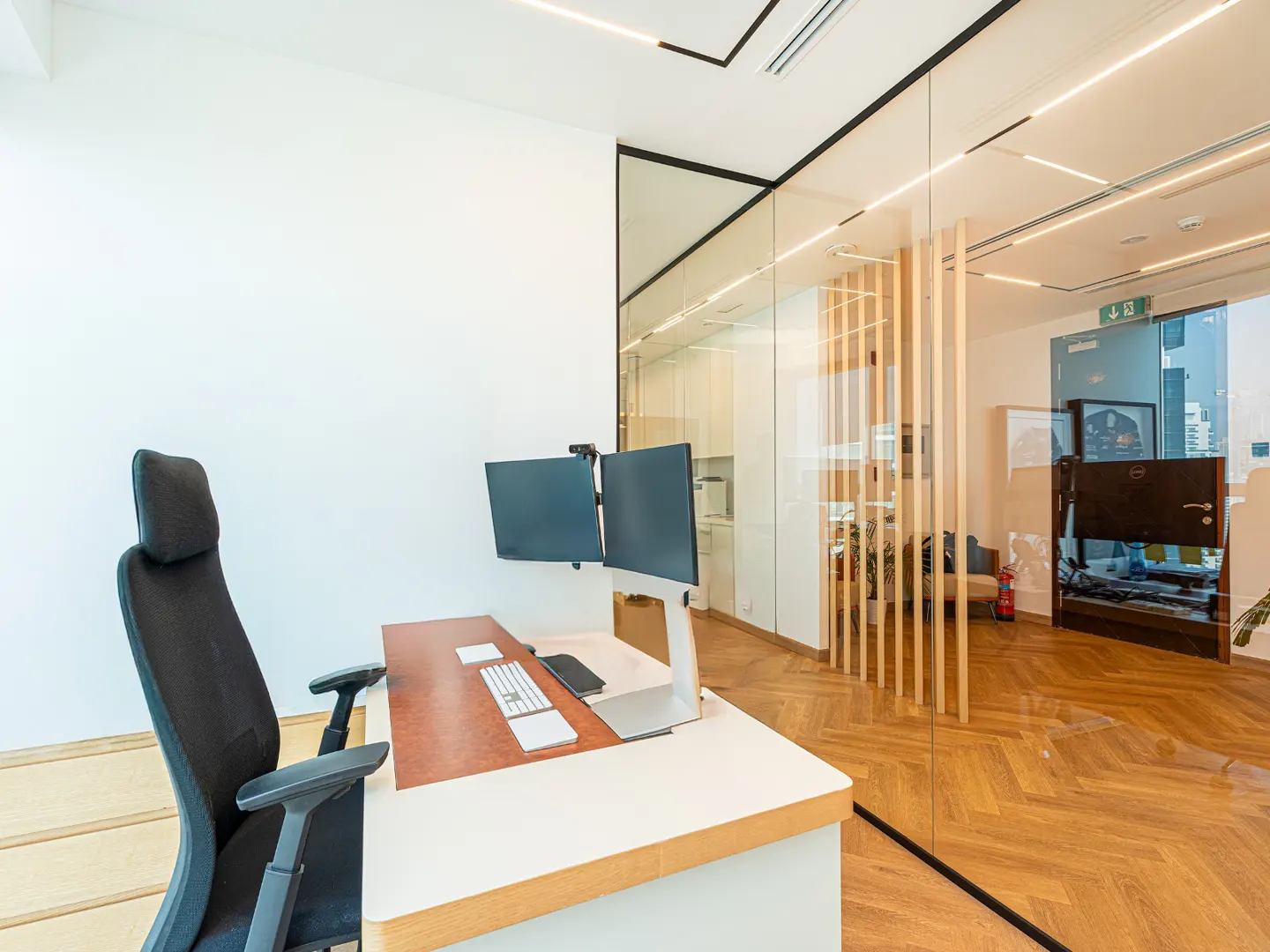 Office interior with a white desk, two monitors, and a black chair. A glass wall separates the office from a room with wood floors.