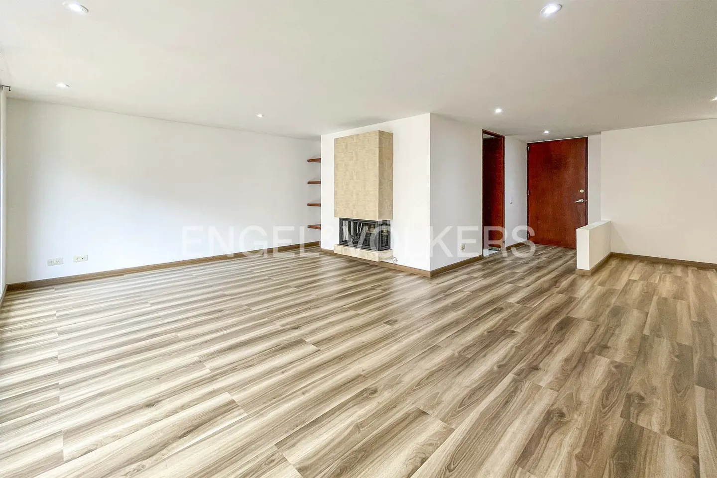 An empty living room with wood-look floors, white walls, and a stone fireplace. A dark wood door is visible.
