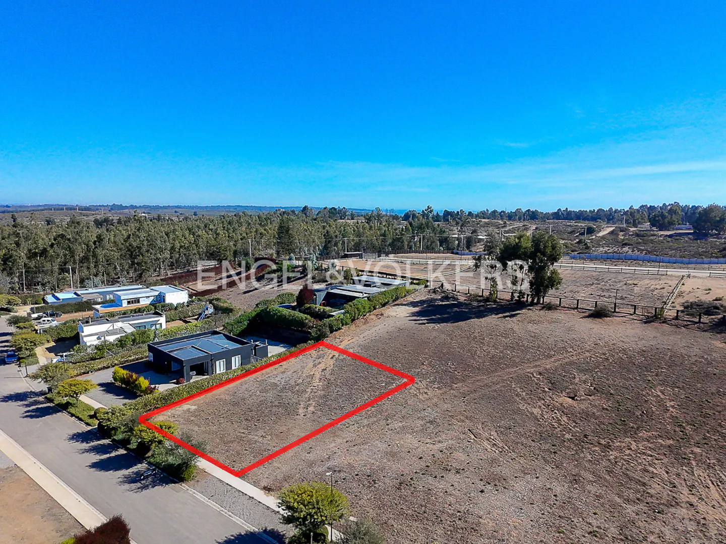 Aerial view of a vacant lot outlined in red, with houses, trees, and a blue sky in the background.