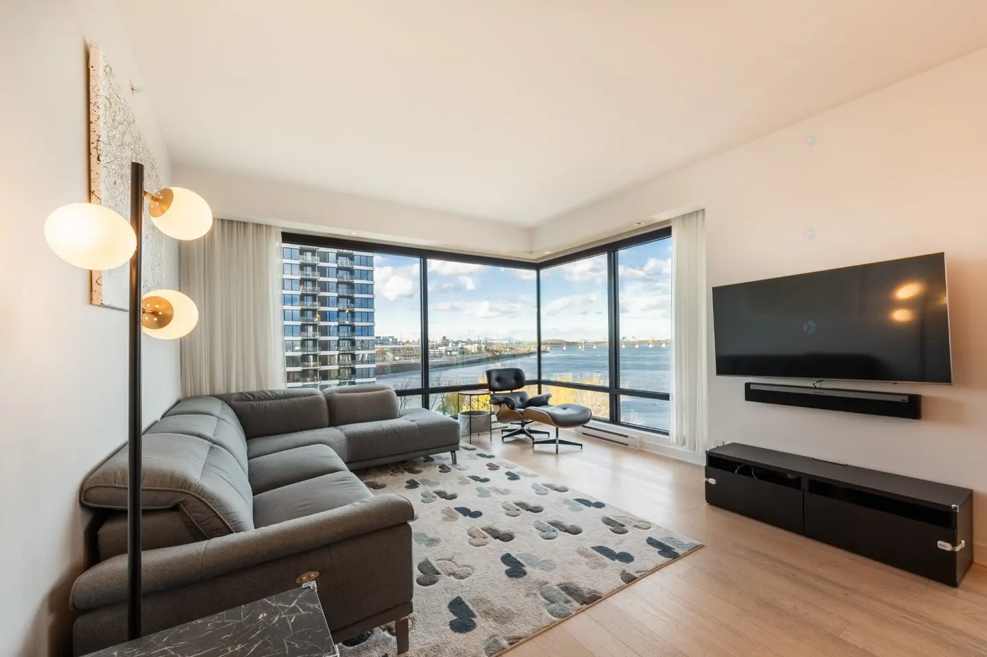 Bright living room with a gray sectional sofa, modern floor lamp, and a large window overlooking a city and river view. A black TV and stand are on the right.