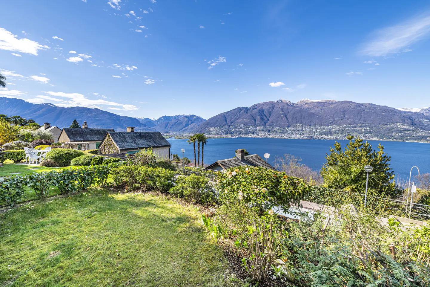 Scenic view of houses with dark roofs, lush green gardens, and a blue lake surrounded by mountains under a clear blue sky.
