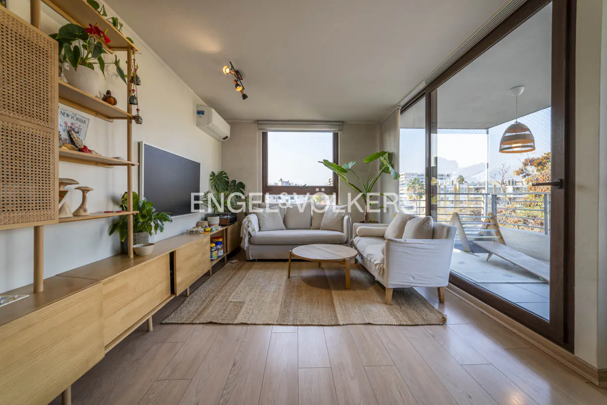 A bright living room with wood floors, a jute rug, and two white sofas. A large window shows a city view. A wooden shelf unit displays plants and decor.