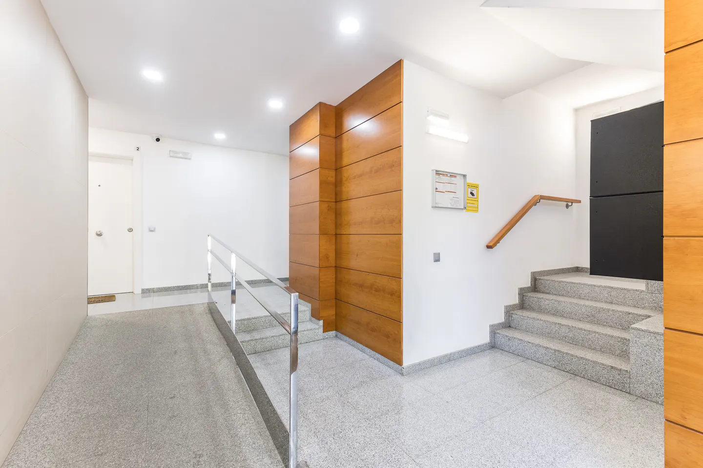 Apartment building hallway with a ramp, stairs, and wood accents. The walls are white, and the floor is speckled gray.