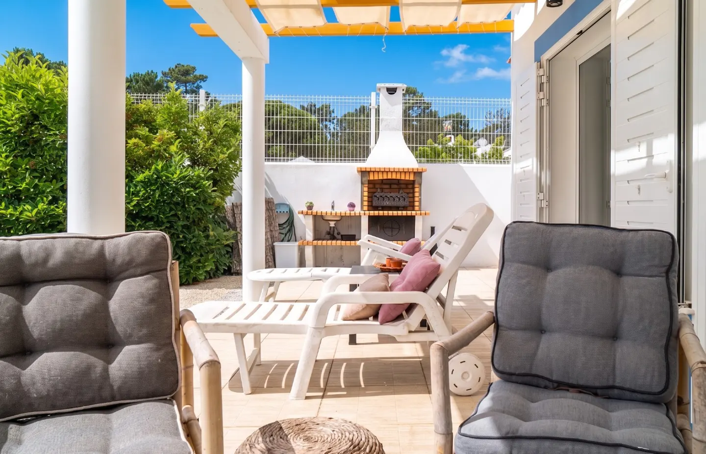 Outdoor patio with gray cushioned chairs, white lounge chairs, and a brick grill. A white fence and green trees are in the background.