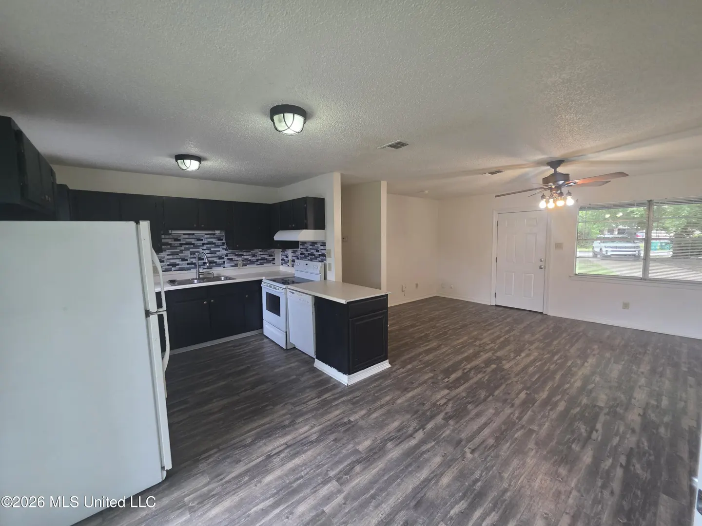 Open-concept kitchen with black cabinets, white appliances, and wood-look flooring, leading to a bright living area.
