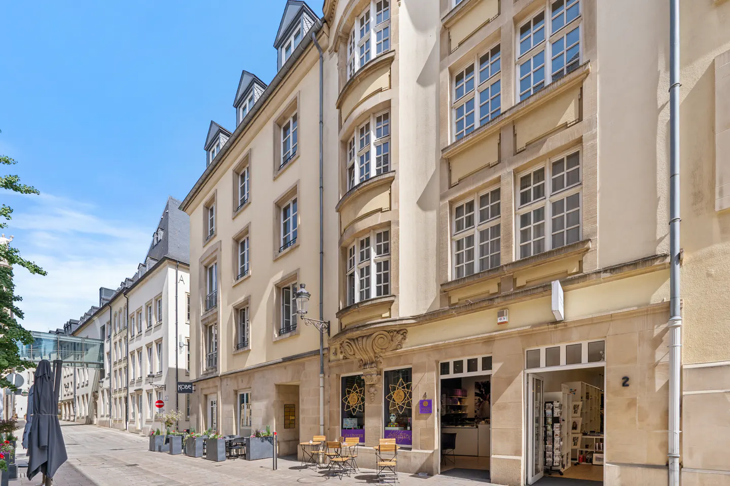A sunny street view of a multi-story cream-colored building with a storefront, outdoor cafe seating, and a clear blue sky.