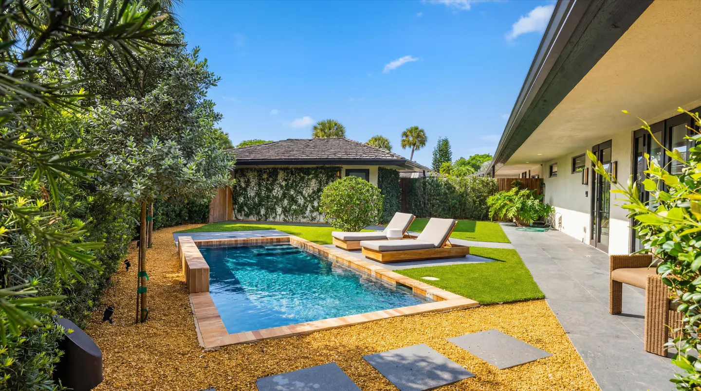 Backyard view of a rectangular pool with two lounge chairs, surrounded by gravel, grass, and lush greenery.