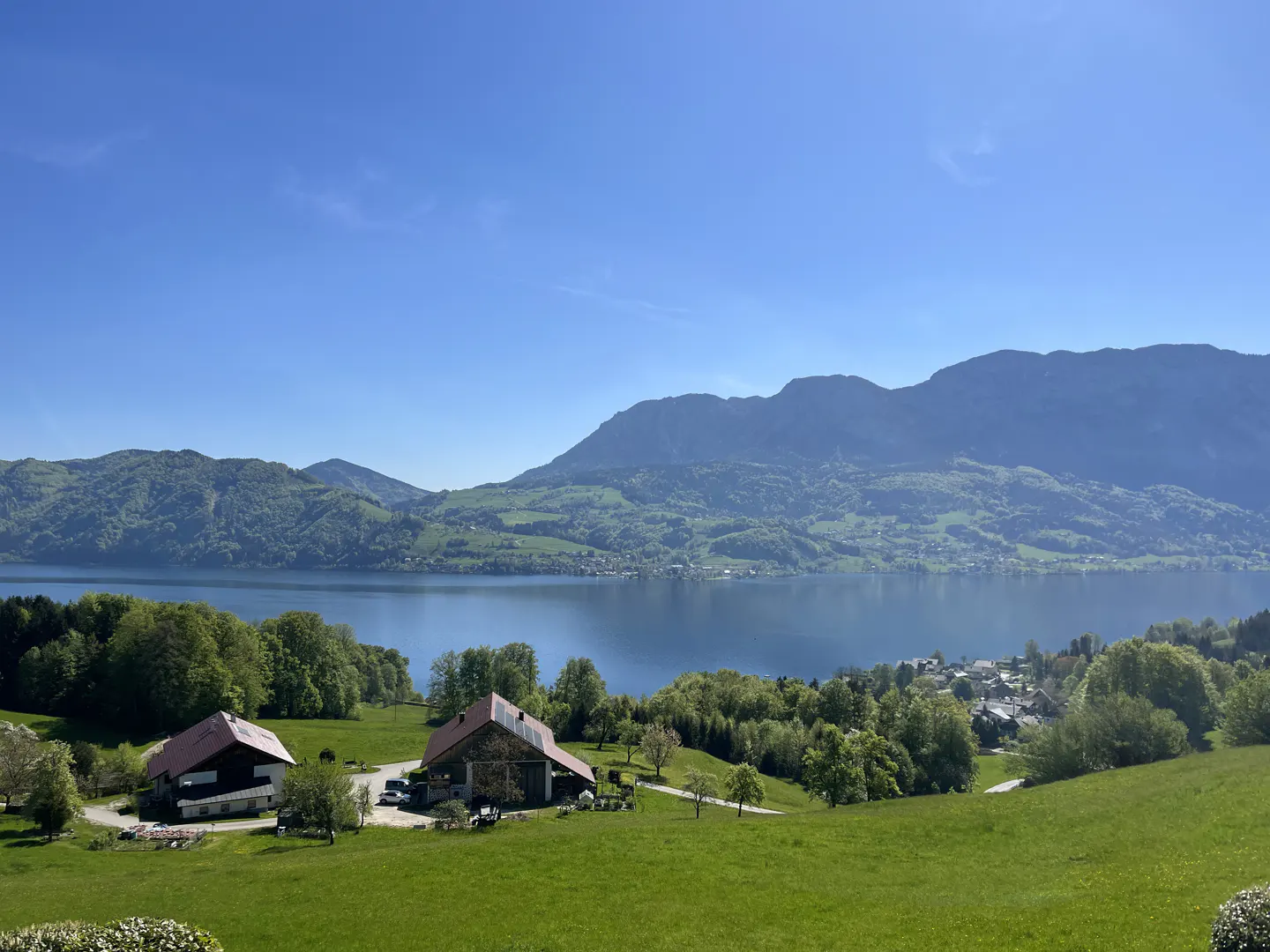 Scenic view of two farmhouses on a green hill overlooking a lake and mountains under a clear blue sky.