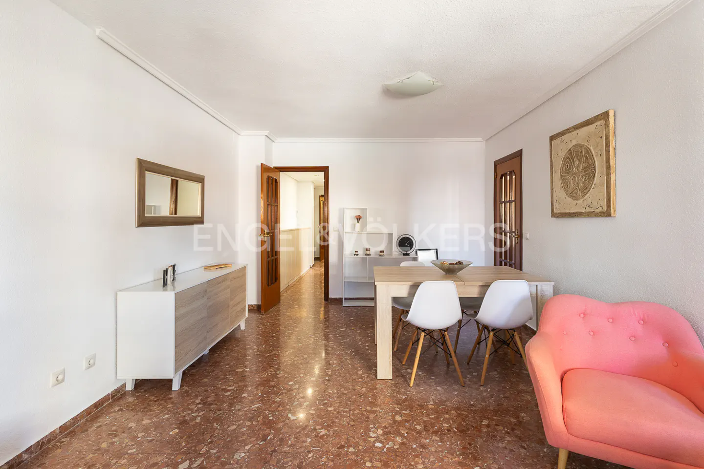 Bright living room with white walls, red terrazzo floor, dining table, pink chair, and modern sideboard.