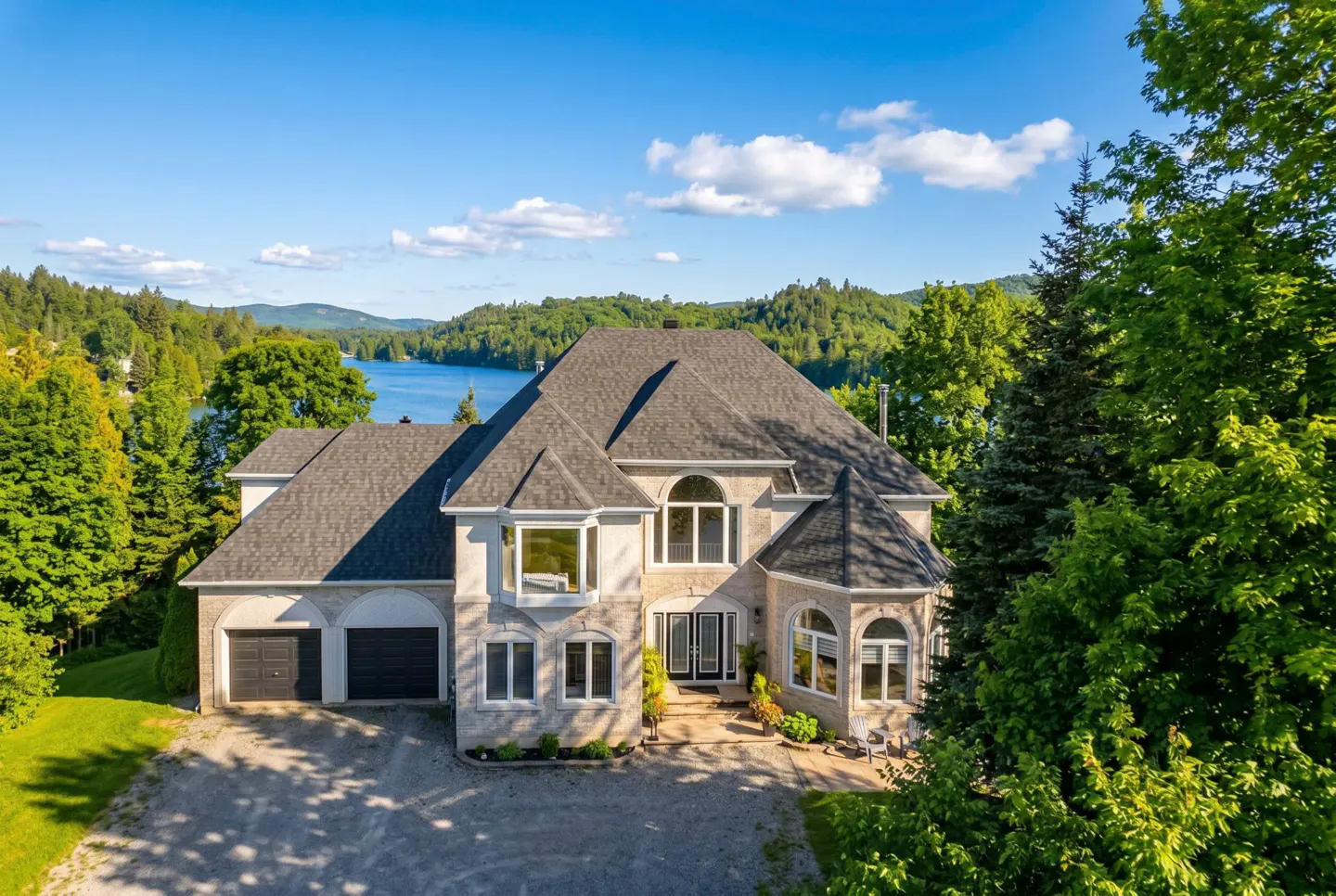 Exterior view of a large, light-stone house with a dark roof, a two-car garage, and a lake in the background.