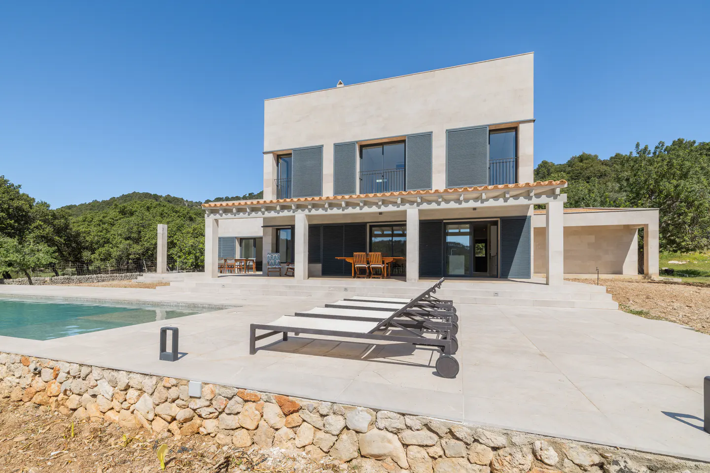 Modern two-story house with a pool and lounge chairs on a sunny day. The house has gray shutters and a covered patio.