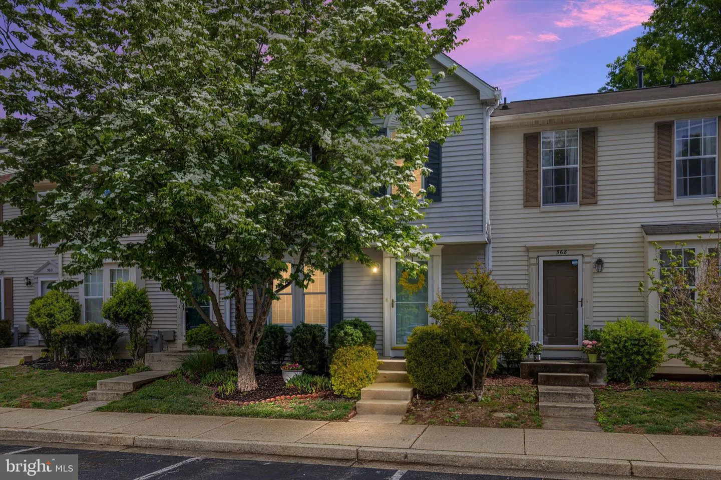 A two-story townhouse with light-blue siding and a large tree with white flowers in front. The sky is a mix of pink and blue.
