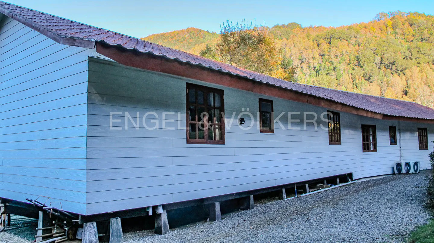 Exterior view of a long, light blue house with a red roof, brown window frames, and a backdrop of forested hills.