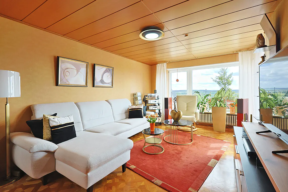 Living room with orange walls and wood ceiling. White sectional sofa, red rug, and large window with a view.