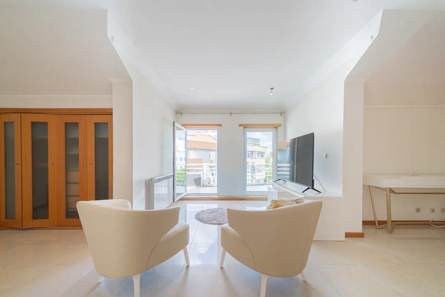 Bright living room with two cream armchairs, a TV, and balcony doors open to a city view. A wooden cabinet is on the left.