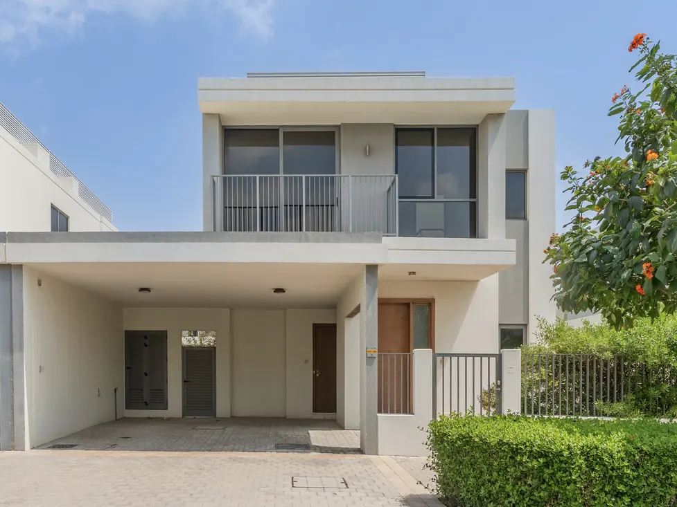 Two-story modern house with a carport, gray trim, and a small balcony. A green hedge and tree with orange flowers are on the right.
