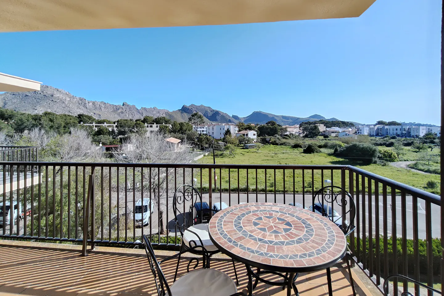 Balcony view with a mosaic table and chairs overlooking green fields, houses, and mountains under a clear blue sky.