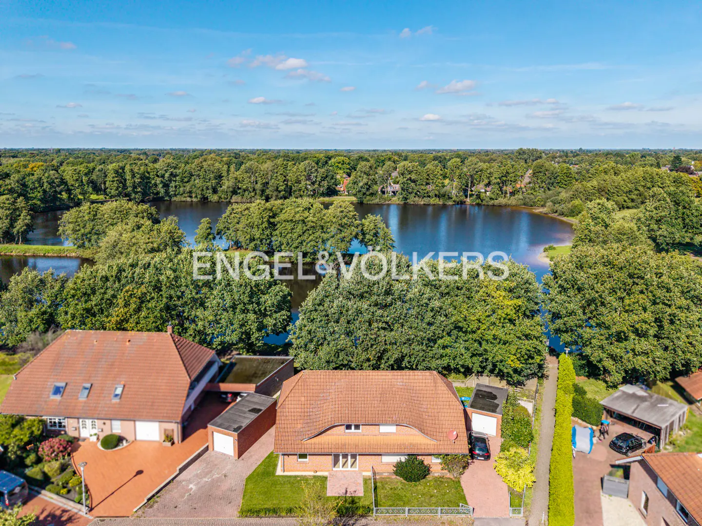 Aerial view of two brick houses with orange tiled roofs, driveways, and garages, situated next to a lake and lush trees.