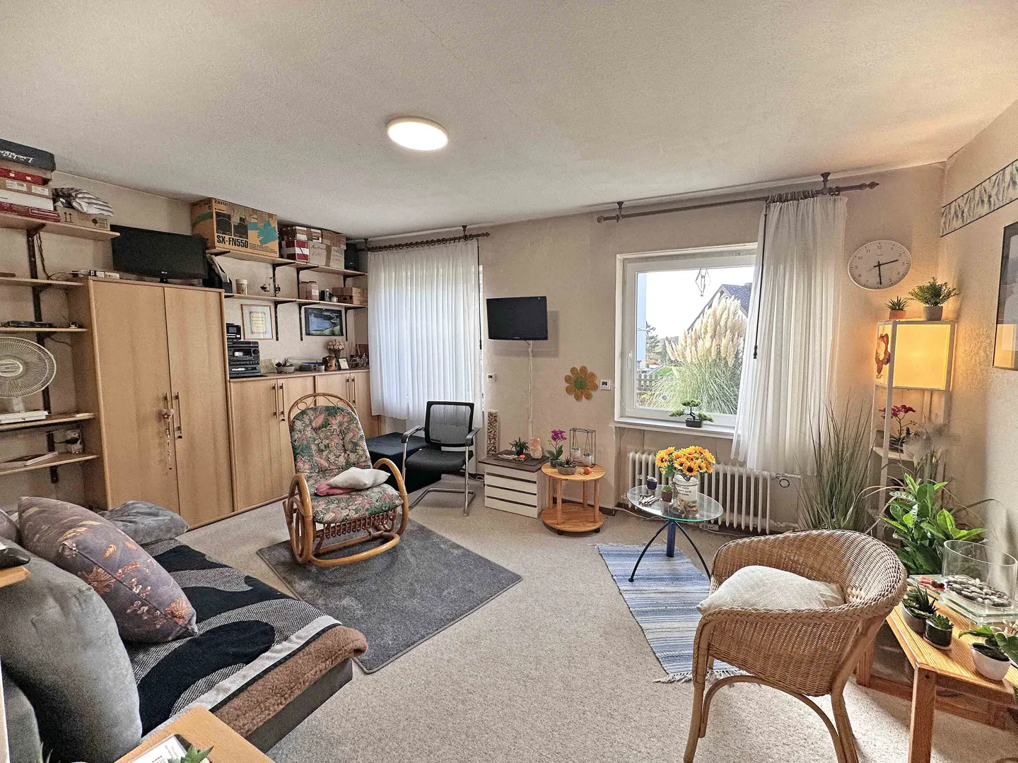 A cozy living room with a sofa, rocking chair, wicker chair, and shelves filled with various items. A window with white curtains lets in natural light.
